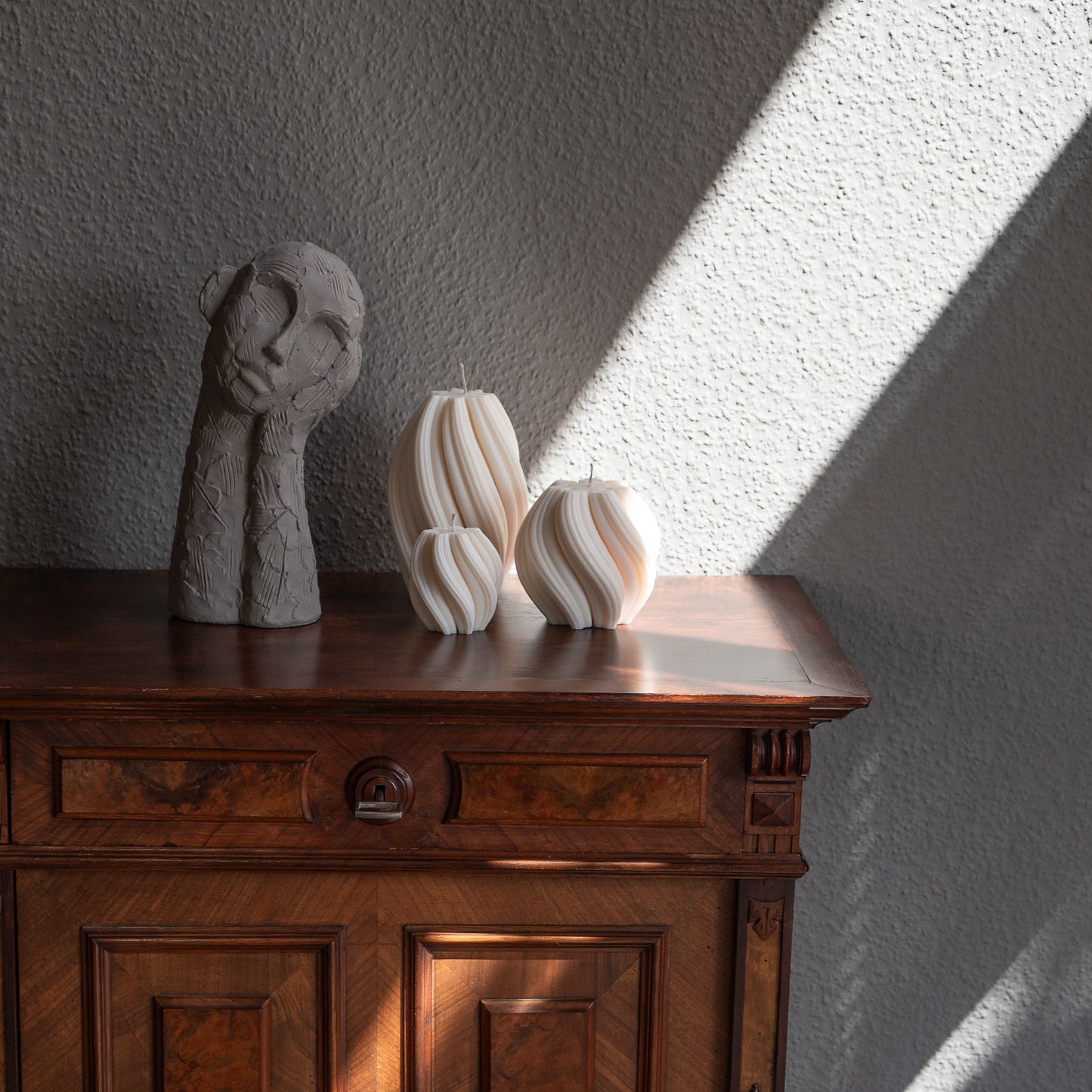 3er Kerzen Set Bøluna in natürlichem Cremeton – skulpturale Designkerzen, Rapswachs, handgefertigt in Hamburg, 100 % pflanzlich & nachhaltig. Studio Z. Stumpenkerzen in Wellenform, geschwungen und bauchig. Dekoriert auf barocker brauner Kommode im Licht der Sonne mit Schattenspielen neben nachdenklicher Betonfigur.