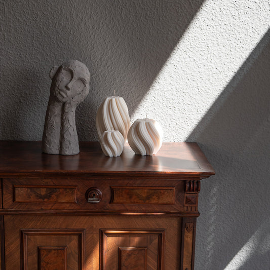 3er Kerzen Set Bøluna in natürlichem Cremeton – skulpturale Designkerzen, Rapswachs, handgefertigt in Hamburg, 100 % pflanzlich & nachhaltig. Studio Z. Stumpenkerzen in Wellenform, geschwungen und bauchig. Dekoriert auf barocker brauner Kommode im Licht der Sonne mit Schattenspielen neben nachdenklicher Betonfigur.