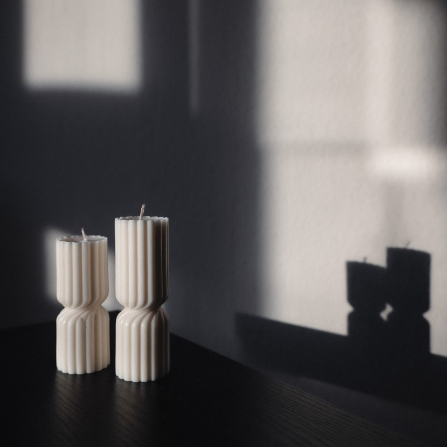 Zweier Kerzen-Set Strøme, mittig gedrehte Stumpenkerze in natürlichem Cremeton. Skulpturale längliche Designkerzen, Rapswachs, handgefertigt in Hamburg, 100 % pflanzlich & nachhaltig. Studio Z. Geriffelte Struktur, dekoriert auf schwarzem Tisch mit rückwärts im Schatten abgebildetem Spiegelbild der Formen.