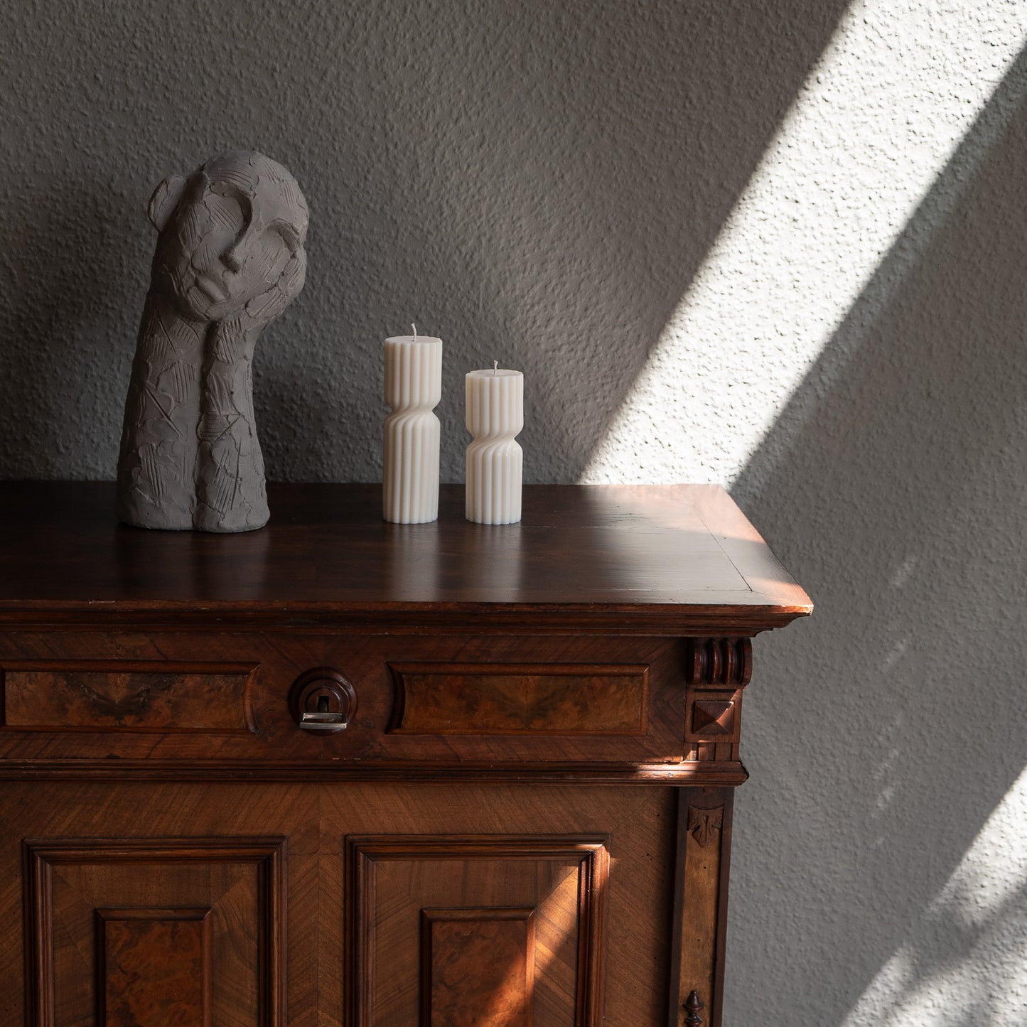 Zweier Kerzen-Set Strøme, mittig gedrehte Stumpenkerze in natürlichem Cremeton. Skulpturale längliche Designkerzen, Rapswachs, handgefertigt in Hamburg, 100 % pflanzlich & nachhaltig. Studio Z. Geriffelte Struktur, dekoriert auf barockem Tisch neben nachdenklicher Betonfigur und einfallendem Sonnenlicht und Schattenspiel.
