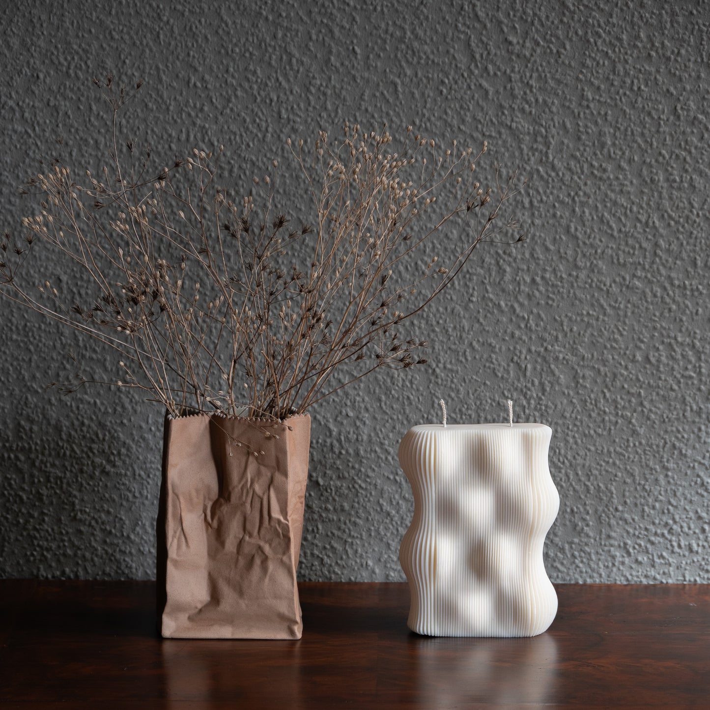 Væg Kerze in natürlichem Cremeton – Designkerzen aus Rapswachs, handgefertigt in Hamburg. 100 % pflanzlich & nachhaltig. Manufaktur Z. Skulpturale Wellenform wie eine horizontale Wachswand. Dekoriert auf barocker Kommode neben brauner Tüten- Vase vor grauer Wand umwunden von Trockenblumen.