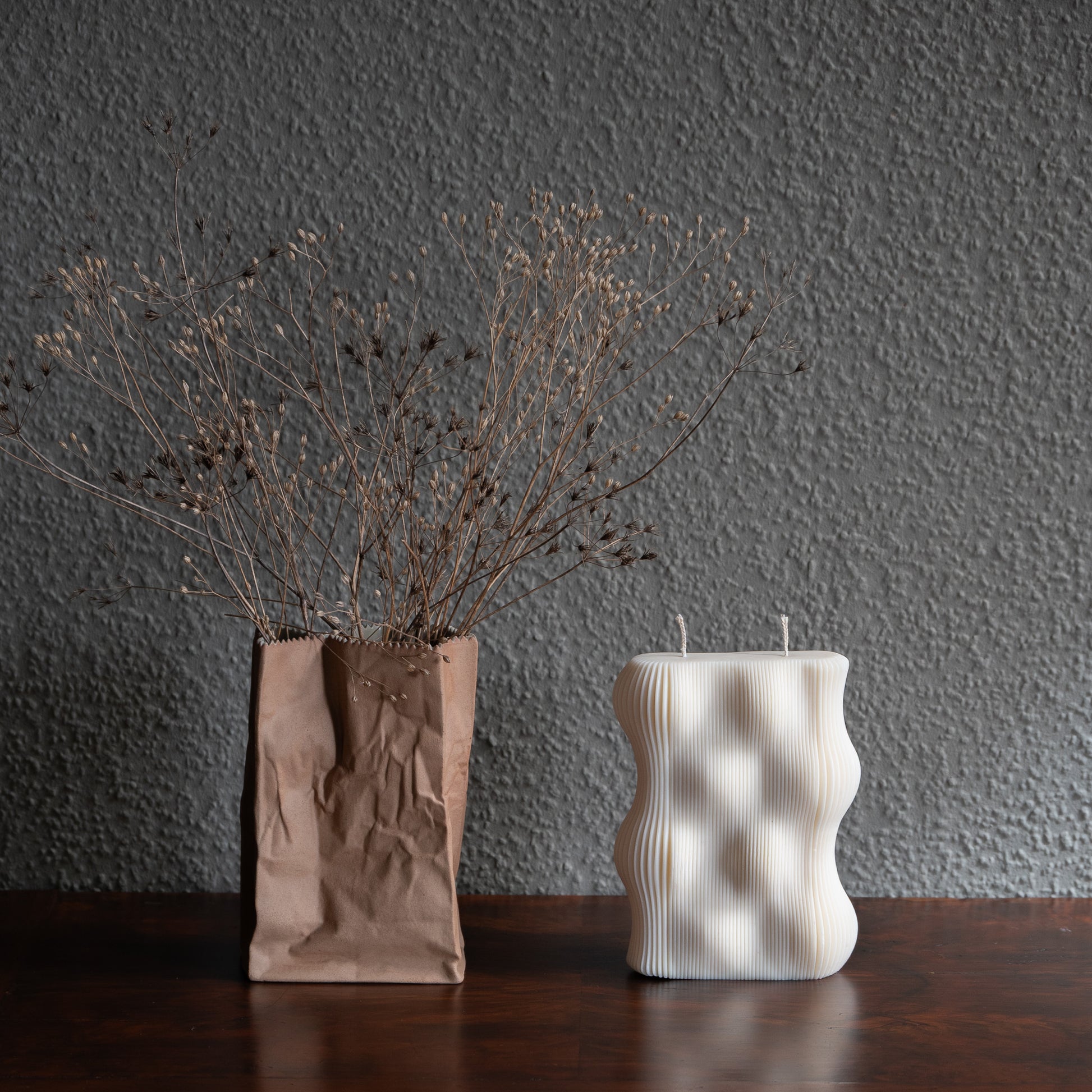 Væg Kerze in natürlichem Cremeton – Designkerzen aus Rapswachs, handgefertigt in Hamburg. 100 % pflanzlich & nachhaltig. Manufaktur Z. Skulpturale Wellenform wie eine horizontale Wachswand. Dekoriert auf barocker Kommode neben brauner Tüten- Vase vor grauer Wand umwunden von Trockenblumen.