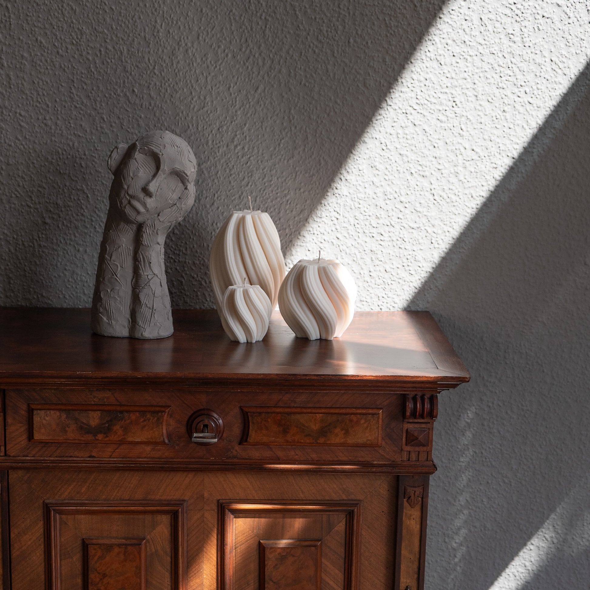 3er Kerzen Set Bøluna in natürlichem Cremeton – skulpturale Designkerzen, Rapswachs, handgefertigt in Hamburg, 100 % pflanzlich & nachhaltig. Studio Z. Stumpenkerzen in Wellenform, geschwungen und bauchig. Dekoriert auf barocker brauner Kommode im Licht der Sonne mit Schattenspielen neben nachdenklicher Betonfigur.