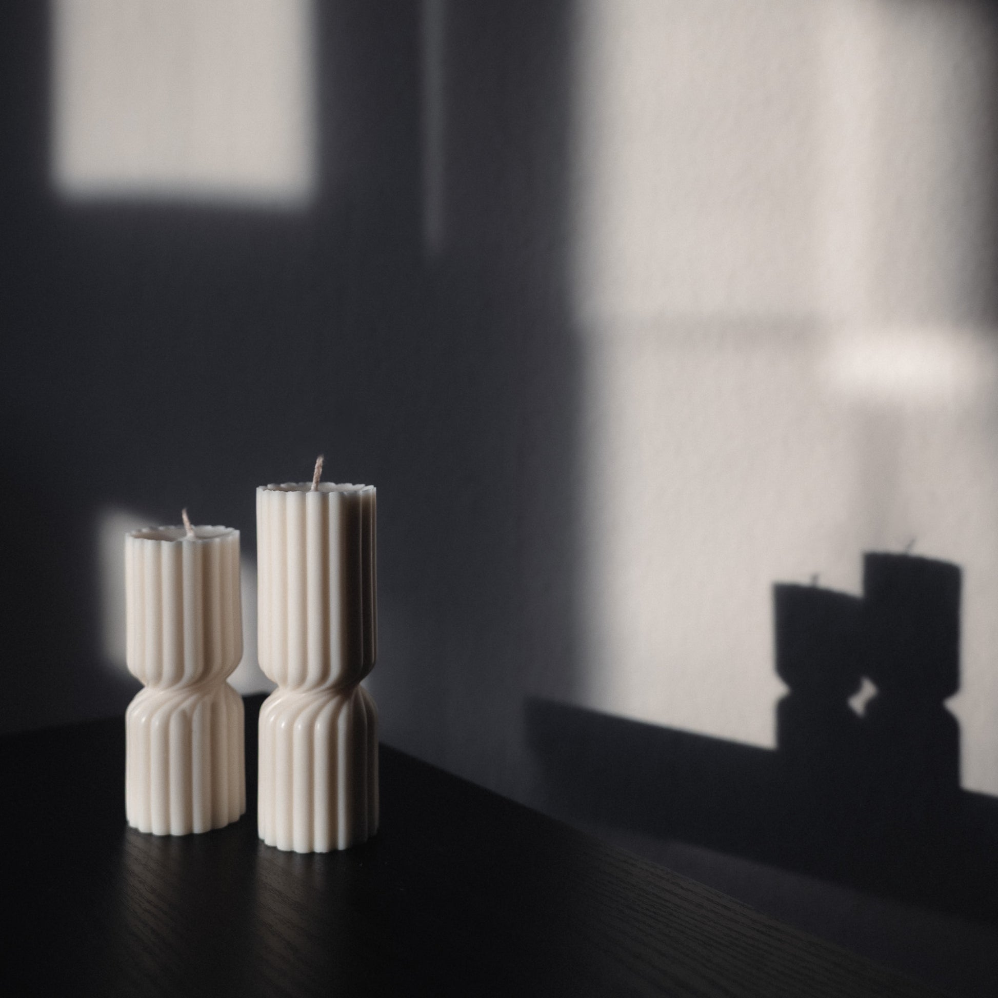 Zweier Kerzen-Set Strøme, mittig gedrehte Stumpenkerze in natürlichem Cremeton. Skulpturale längliche Designkerzen, Rapswachs, handgefertigt in Hamburg, 100 % pflanzlich & nachhaltig. Studio Z. Geriffelte Struktur, dekoriert auf schwarzem Tisch mit rückwärts im Schatten abgebildetem Spiegelbild der Formen.